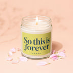 Candle with a lime green label reading "So this is forever" on a pink surface.