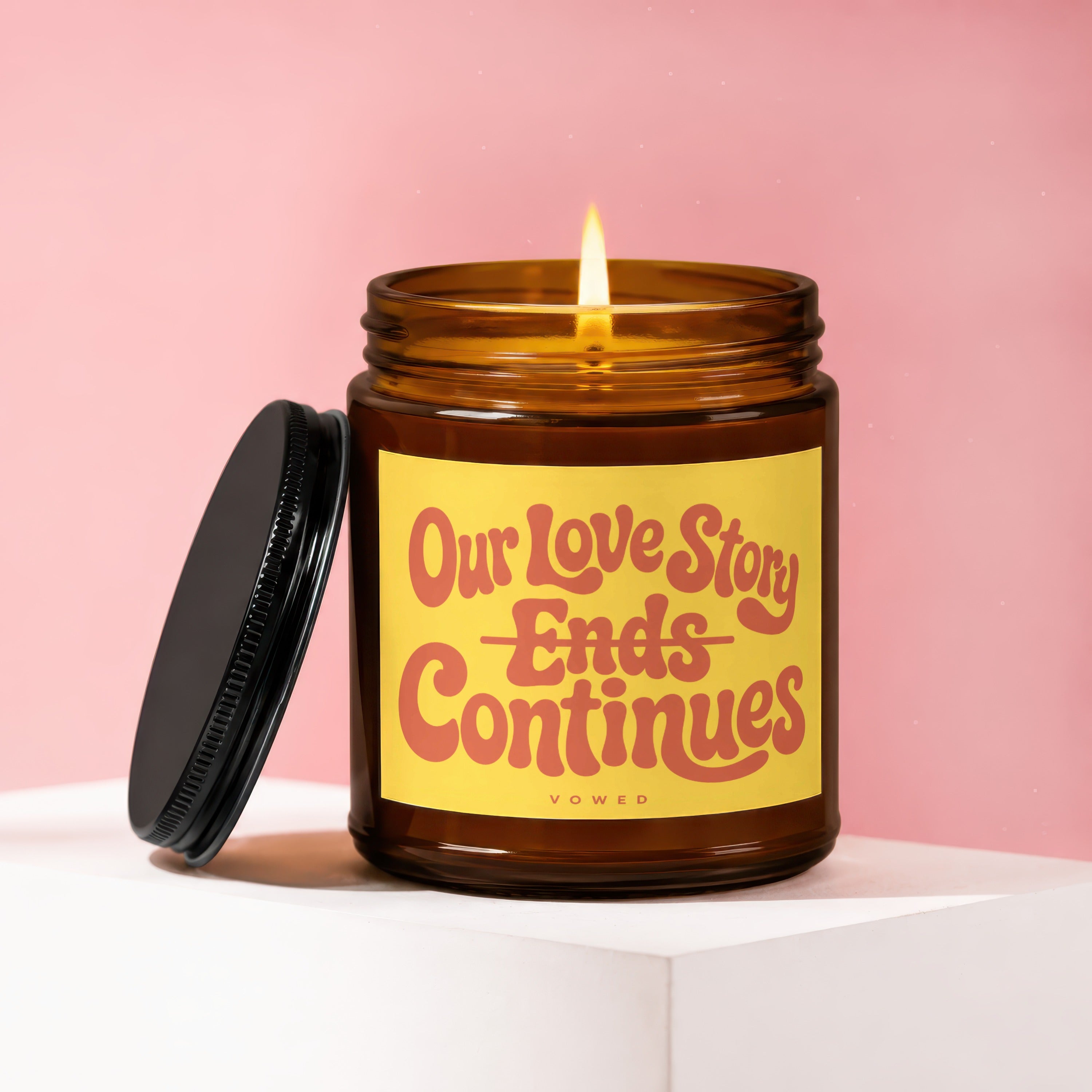 Amber glass candle with lit flame and lid beside it, text reads "Our Love Story Ends Continues".