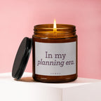 Amber jar candle with lit wick, lid off, on white pedestal. Text: In my planning era.