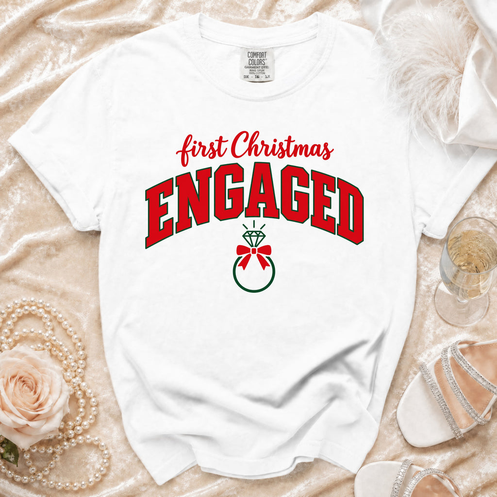 A white t-shirt featuring the text First Christmas Engaged in red and green lettering with a ring graphic.
