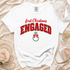 A white t-shirt featuring the text First Christmas Engaged in red and green lettering with a ring graphic.