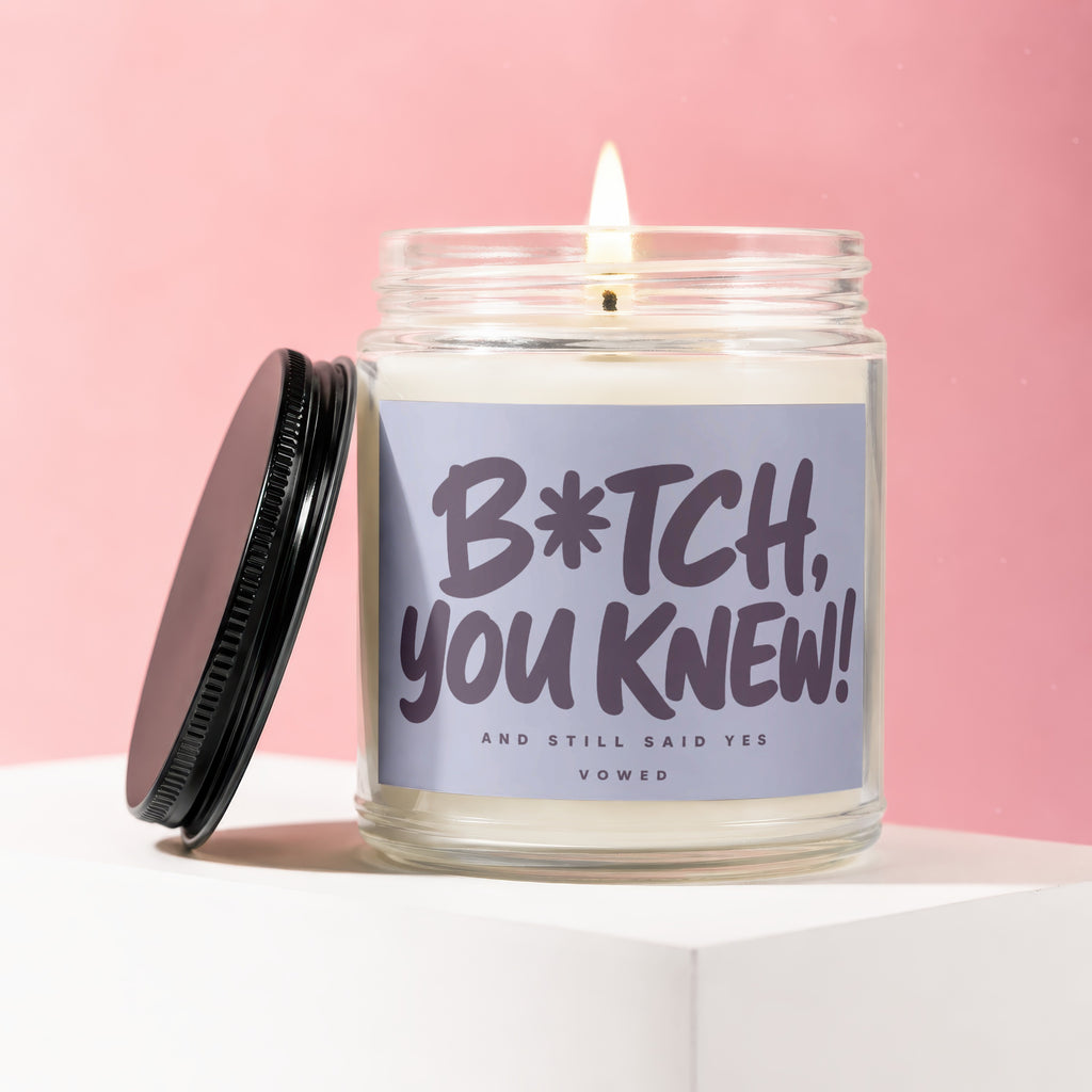 Glass candle with lit wick, black lid, and label reading "B*tch, you knew! And still said yes VOWED".