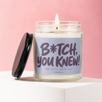 Glass candle with lit wick, black lid, and label reading "B*tch, you knew! And still said yes VOWED".