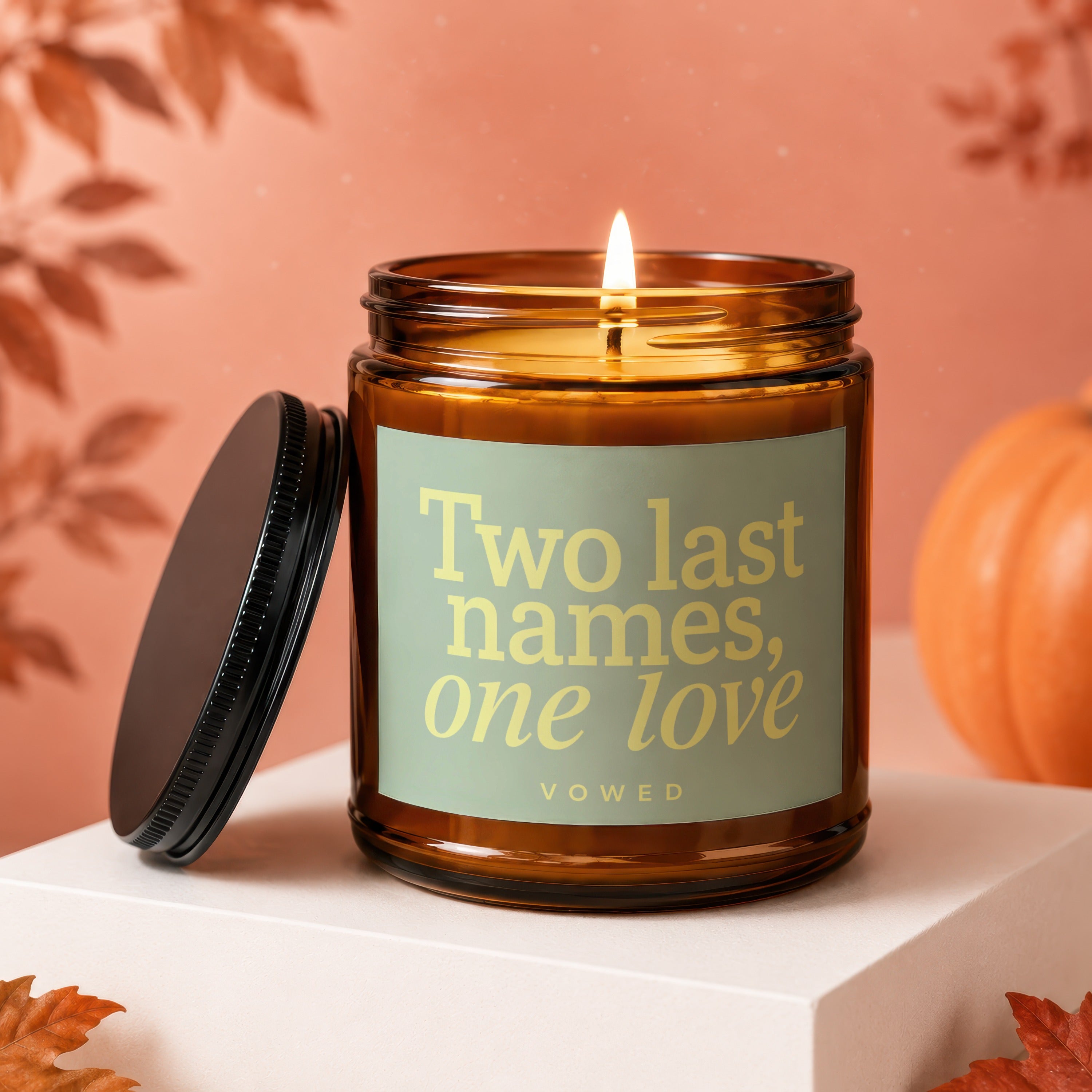 Amber candle with lit wick, lid off, and text "Two last names, one love VOWED".
