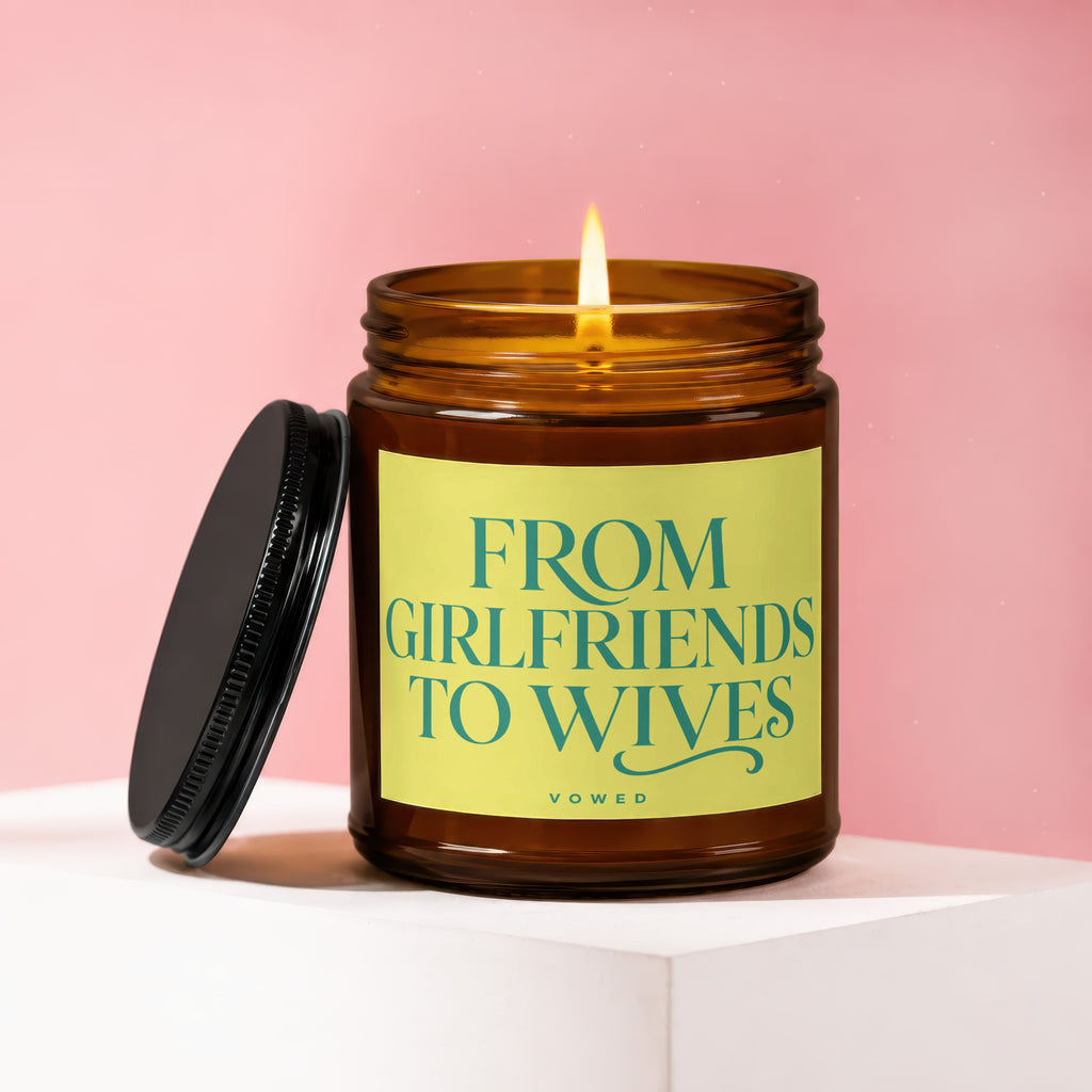 Amber glass candle with lit flame and black lid, reads "FROM GIRLFRIENDS TO WIVES".