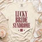 A cream-colored t-shirt with maroon text reading "Lucky Bride Syndrome" above a four-leaf clover.