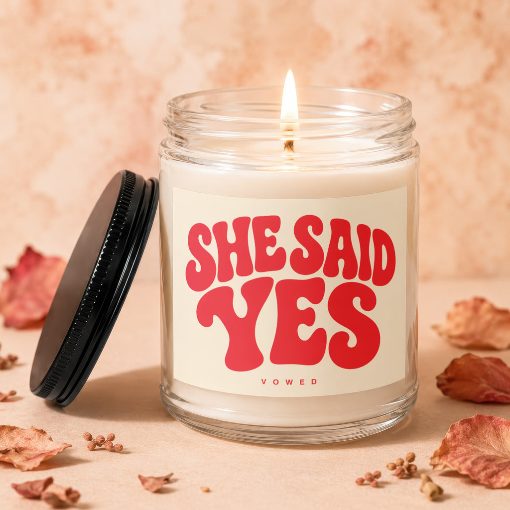 Clear glass candle with a lit wick and red text reading "SHE SAID YES VOWED".