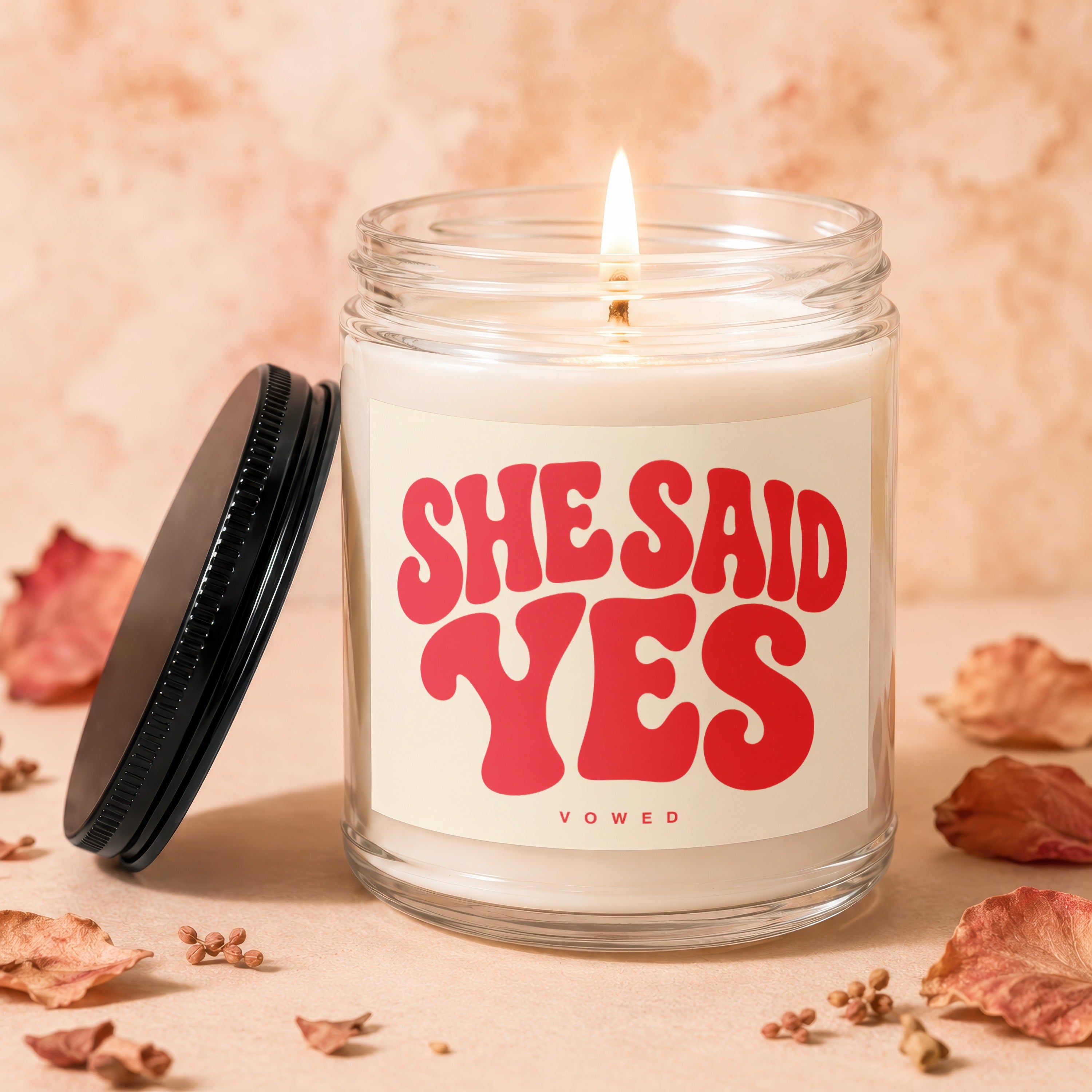 Clear glass candle with a lit wick and red text reading "SHE SAID YES VOWED".