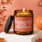 Amber candle with lit wick, lid beside, reading "Engaged & Thrilled VOWED".