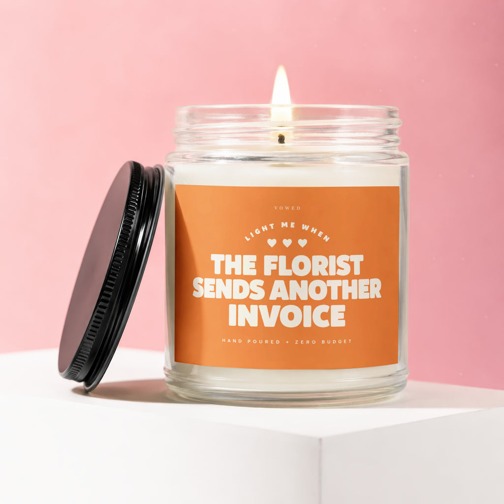 Scented candle in glass jar with orange label reading "THE FLORIST SENDS ANOTHER INVOICE".