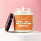 Scented candle in glass jar with orange label reading "THE FLORIST SENDS ANOTHER INVOICE".