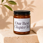 Amber jar candle with black lid, label reads "Our Best Chapter Yet".