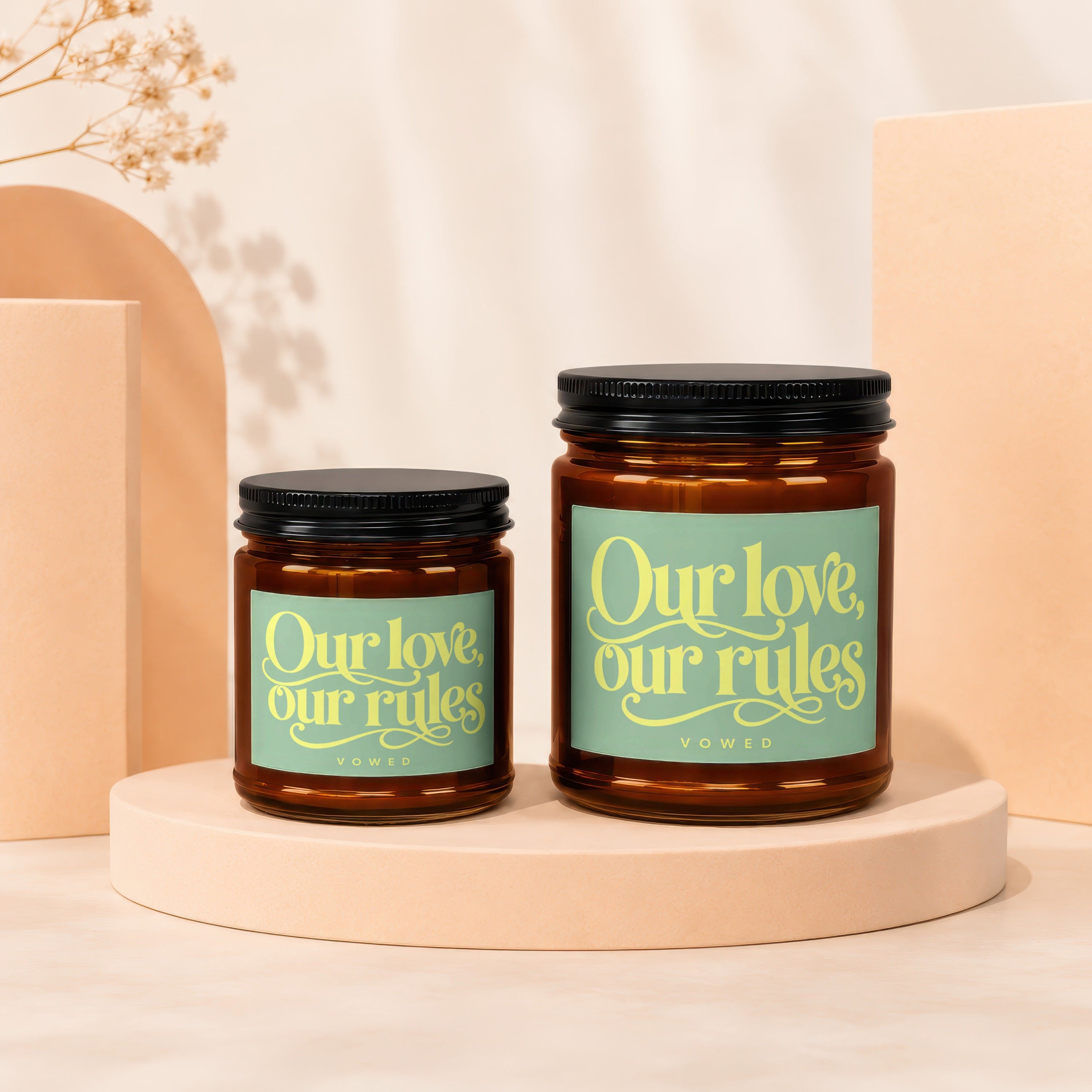 Two amber glass candles with black lids, one larger than the other, sit on a cream-colored pedestal.