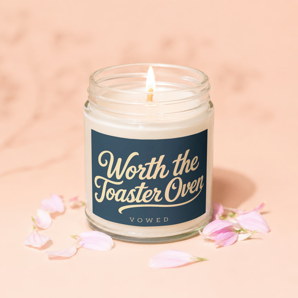 Lit candle in a jar with navy label reading "Worth the Toaster Oven VOWED".