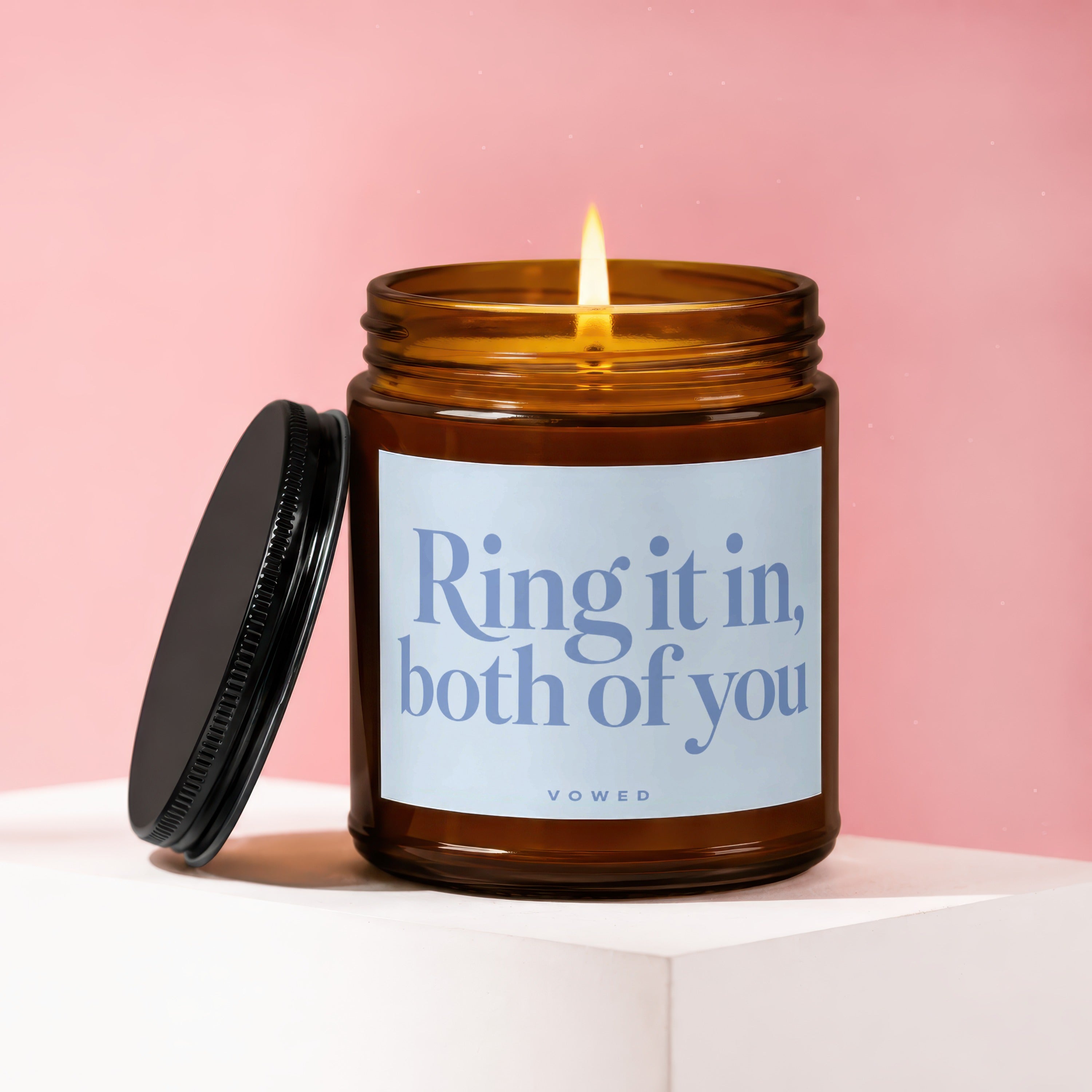 Amber glass candle with a lit wick and black lid, reads "Ring it in, both of you vowed."