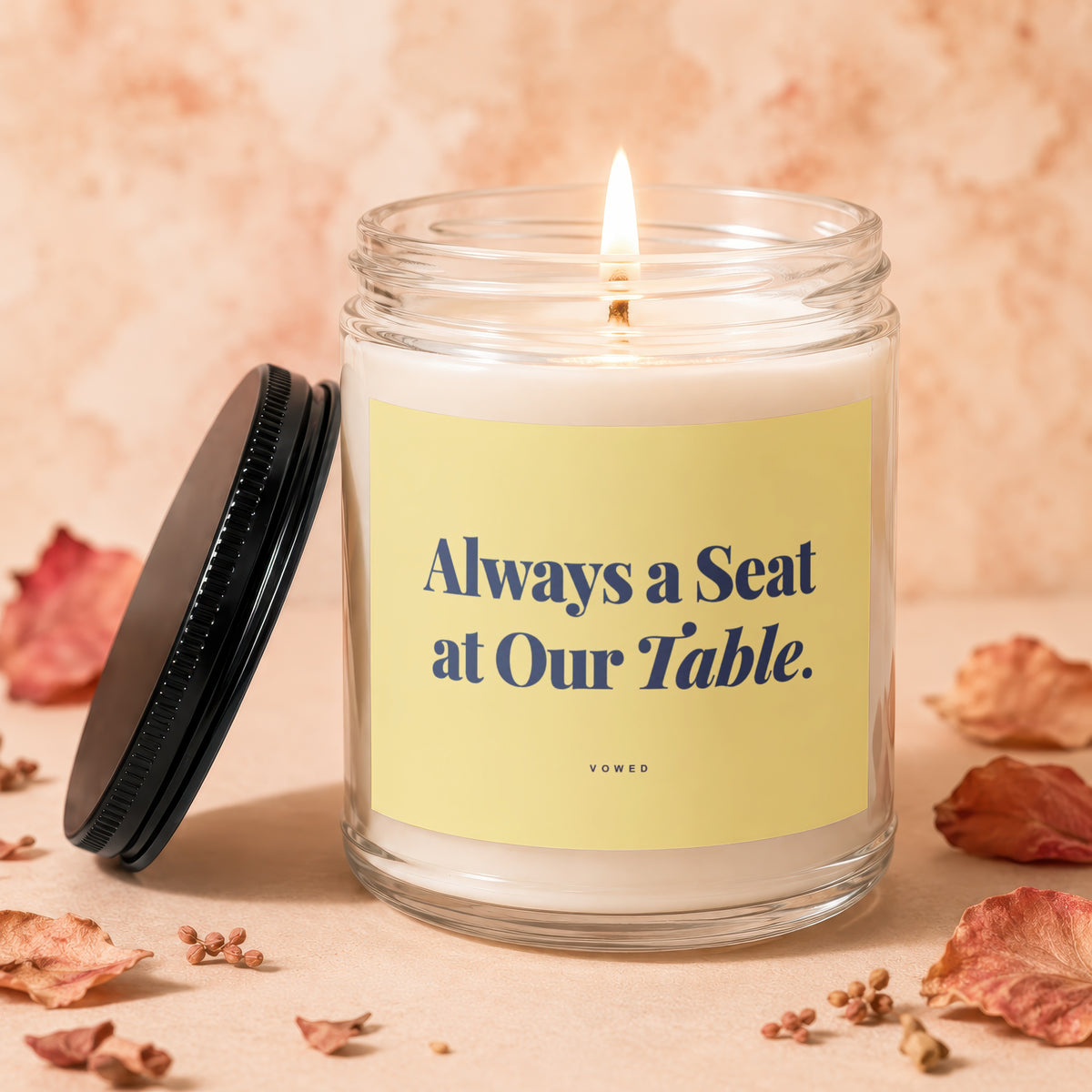 Jar candle with lit wick, yellow label reading "Always a Seat at Our Table. VOWED."