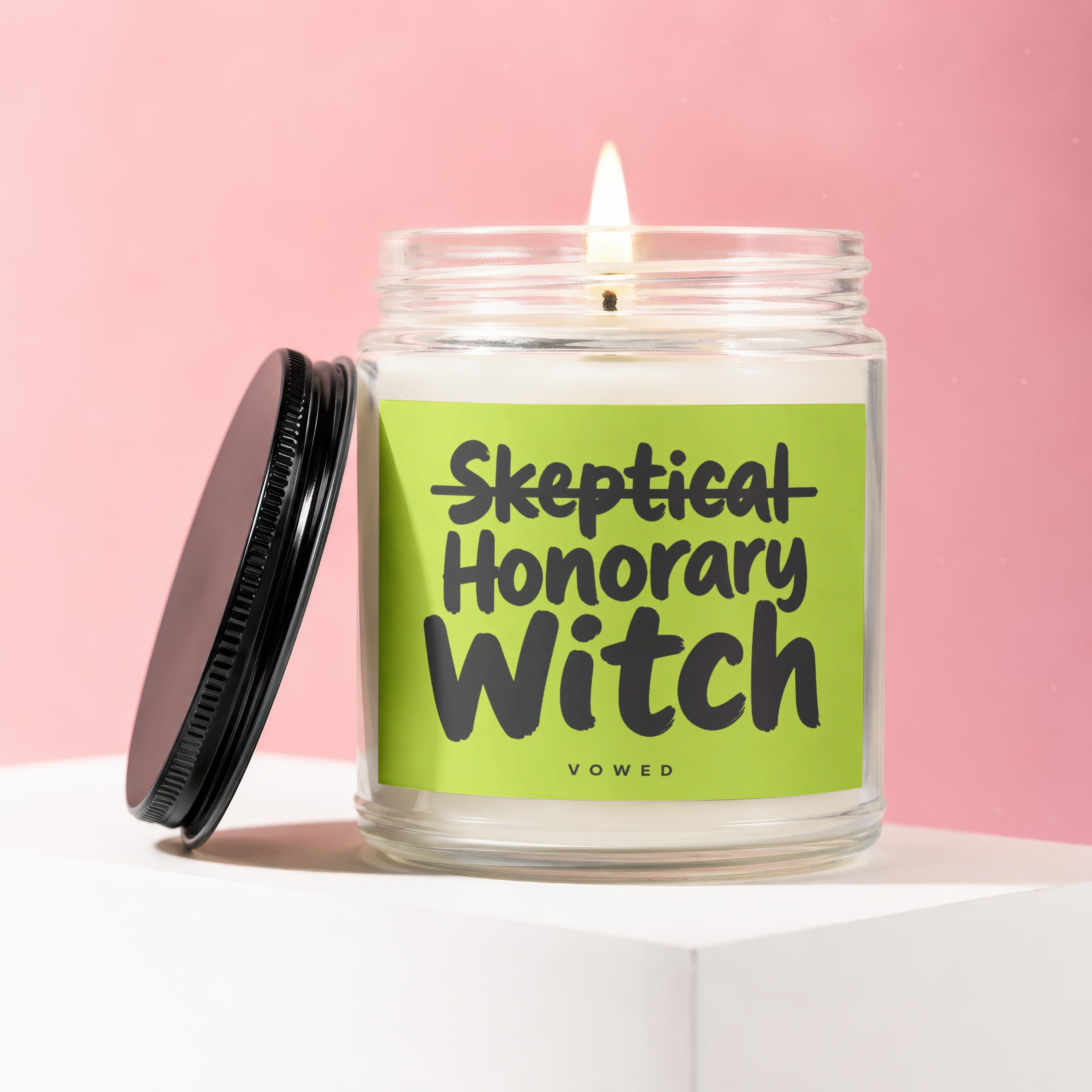 Glass jar candle with lit wick and label reading "Skeptical Honorary Witch".