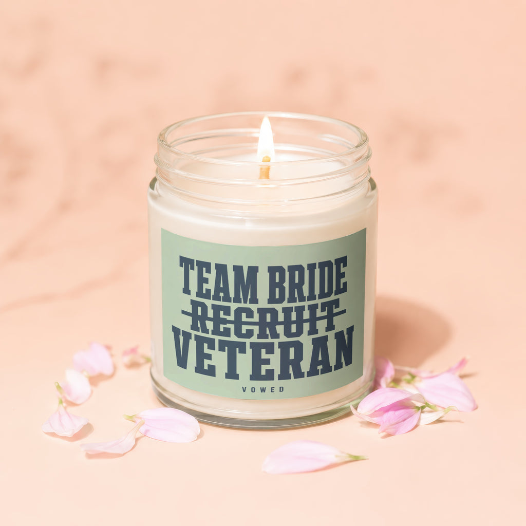 Glass jar candle with lit wick, labeled "Team Bride Recruit Veteran Vowed."