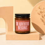 Amber jar candle with "THE ADVENTURE BEGINS" text on a peach backdrop.