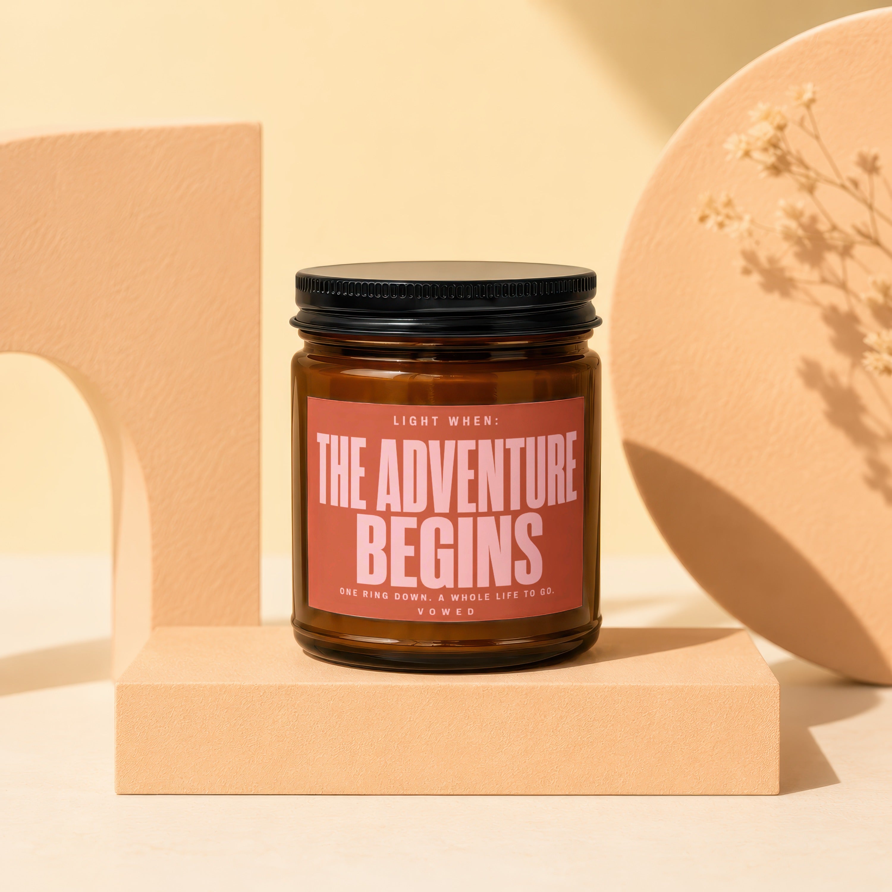 Amber jar candle with "THE ADVENTURE BEGINS" text on a peach backdrop.