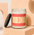 Jar candle with red label reading "BRIDESMAID" and "VOWED", black lid nearby.