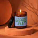 Lit amber glass candle with label reading "IT'S GIVING ENGAGED AF".