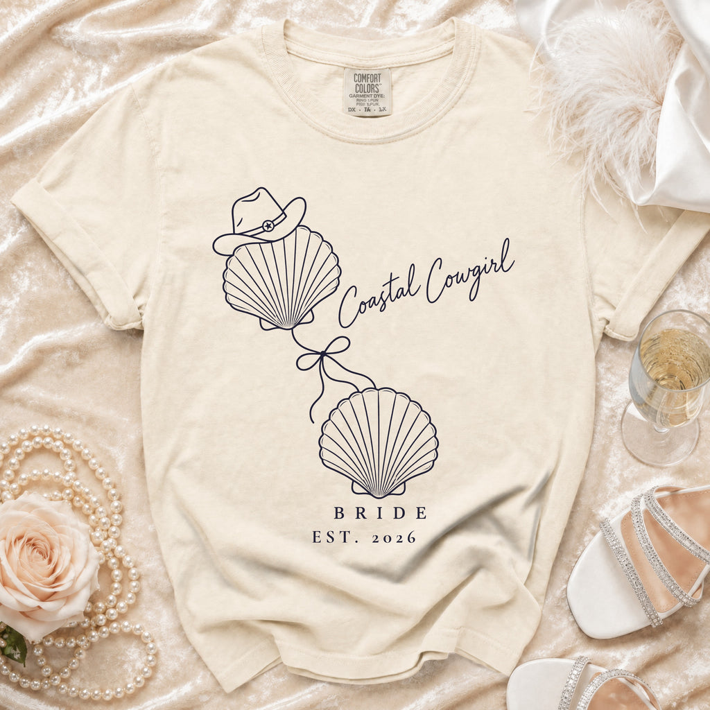 A cream-colored t-shirt with a blue graphic of two seashells, a cowboy hat, and the text "Coastal Cowgirl Bride Est. 2026."