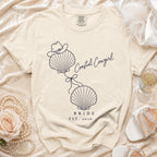 A cream-colored t-shirt with a blue graphic of two seashells, a cowboy hat, and the text "Coastal Cowgirl Bride Est. 2026."