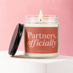 Glass candle with lit wick and black lid, labeled "Partners, officially VOWED".