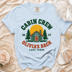 Light blue crewneck t-shirt featuring a green and orange graphic with a cabin, trees, sun, and text that reads CABIN CREW OLIVIA'S BACH LAKE TAHOE.