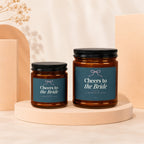 Two amber glass candles with "Cheers to the Bride" labels on a neutral background.