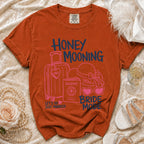 Orange t-shirt with "Honey Mooning Bride Mode" graphic, suitcase, passport, hat, glasses.