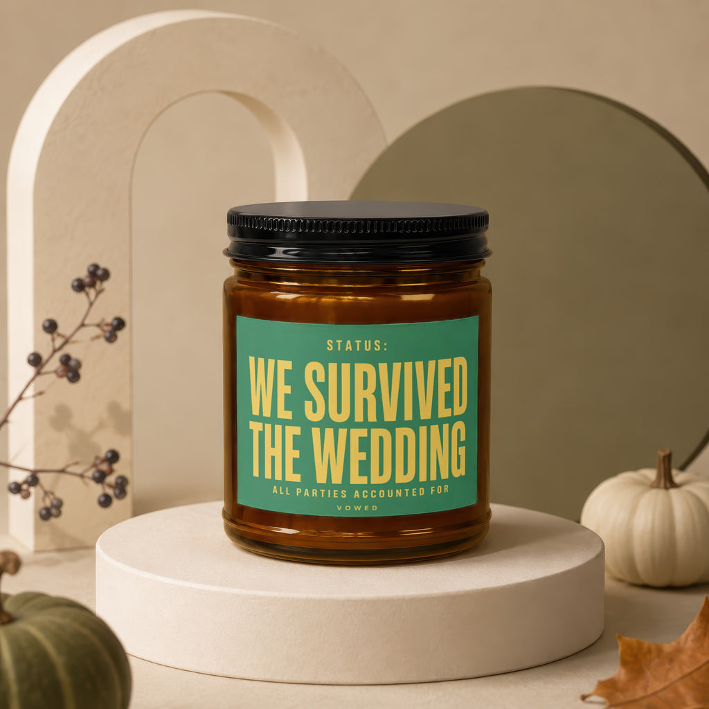 Amber candle in glass jar with black lid and green label reading "WE SURVIVED THE WEDDING".
