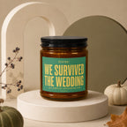 Amber candle in glass jar with black lid and green label reading "WE SURVIVED THE WEDDING".