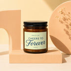 Brown glass candle with "Cheers to Forever" label, on a peach block.