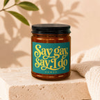Amber jar candle with black lid, features teal label with yellow text "Say gay Say I do VOWED".