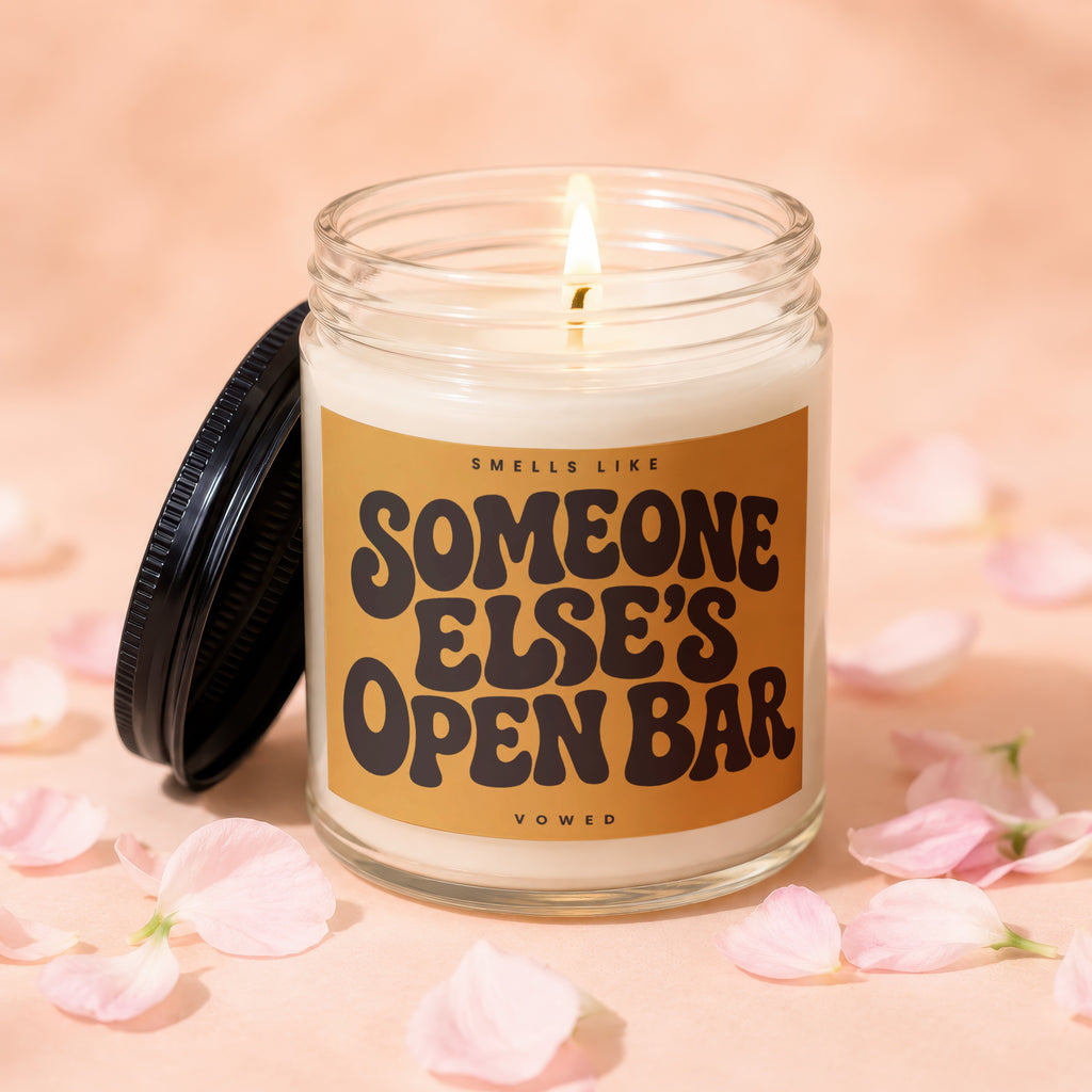 Scented candle with "Smells Like Someone Else's Open Bar" on label surrounded by pink petals.