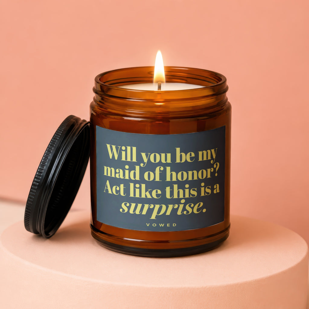 Amber jar candle with lit wick and lid asks "Will you be my maid of honor? Act like this is a surprise."