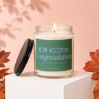 A lit candle in a glass jar sits on a white block, featuring a label with text.