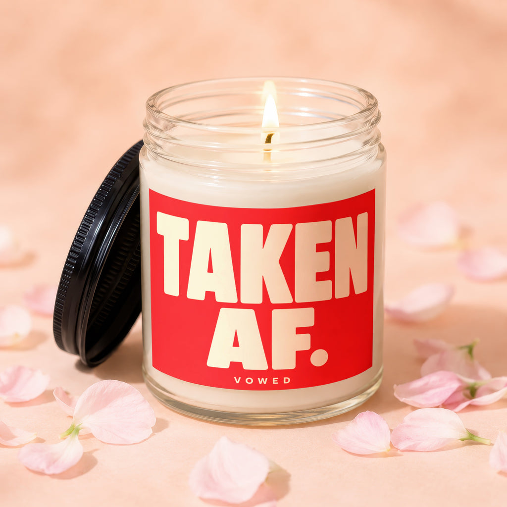 Glass candle lit with burning wick, red label reads "TAKEN AF. VOWED."