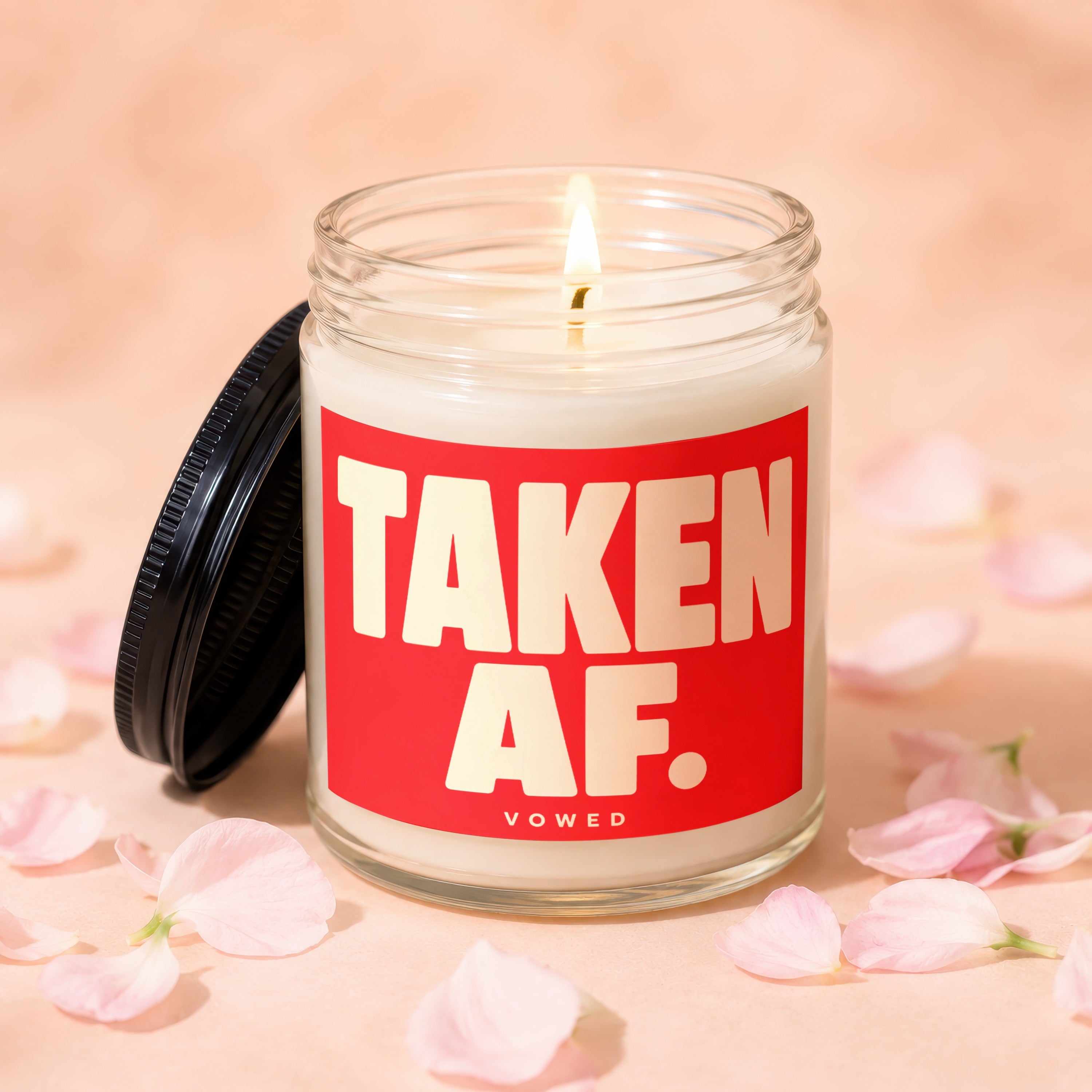 Glass candle lit with burning wick, red label reads "TAKEN AF. VOWED."