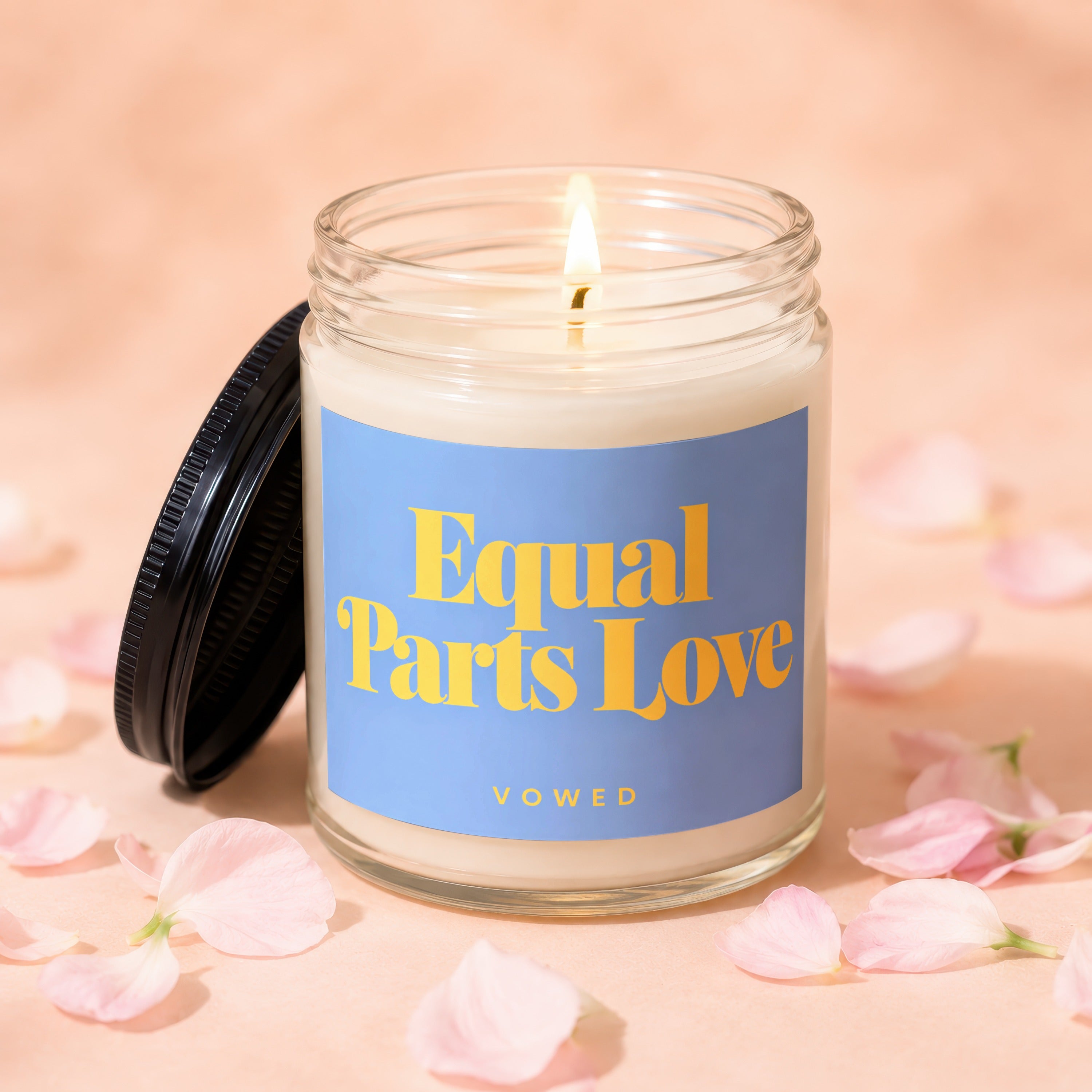 Glass candle with lit wick and blue label reading "Equal Parts Love VOWED".