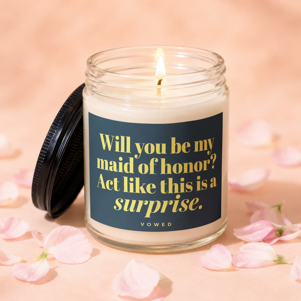 Glass candle with lit wick featuring "Will you be my maid of honor? Act like this is a surprise." text.