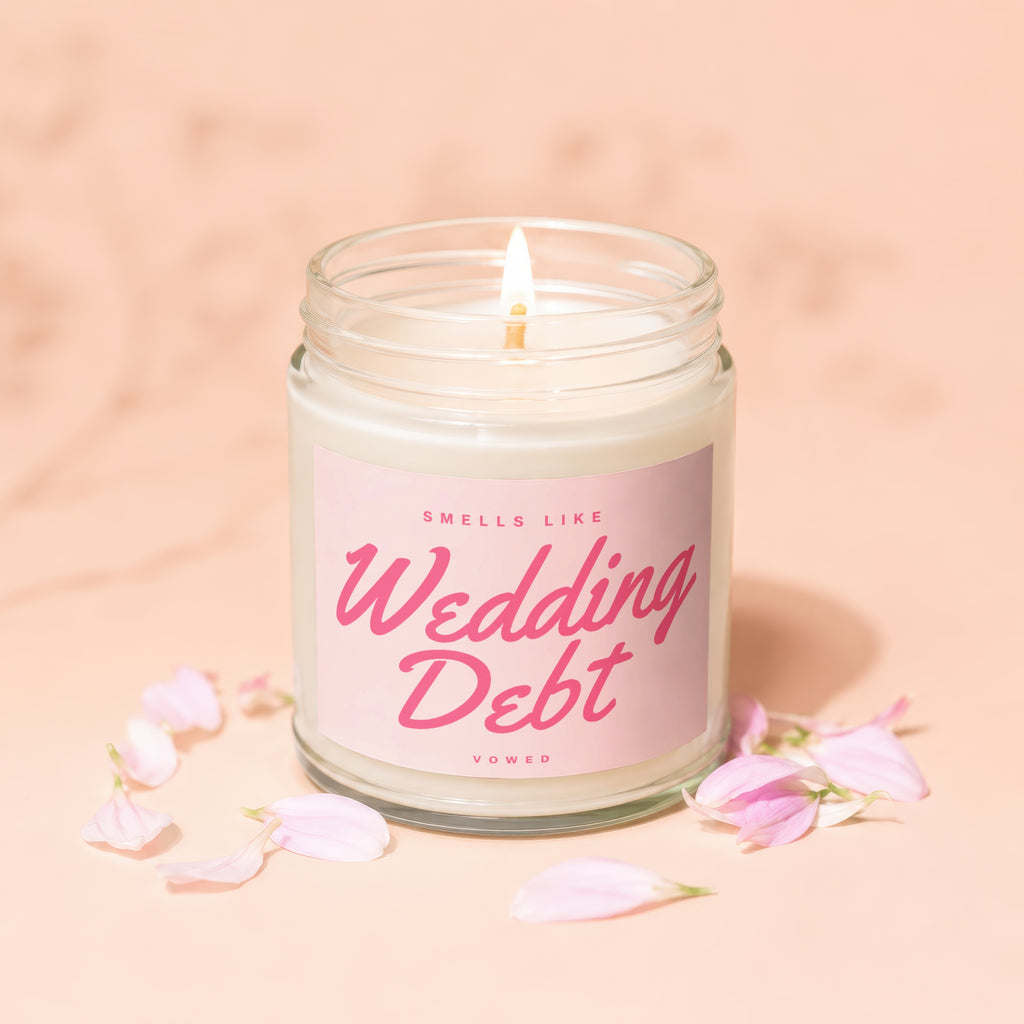 Lit candle in glass jar with "Smells Like Wedding Debt" label surrounded by pink petals.
