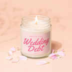 Lit candle in glass jar with "Smells Like Wedding Debt" label surrounded by pink petals.