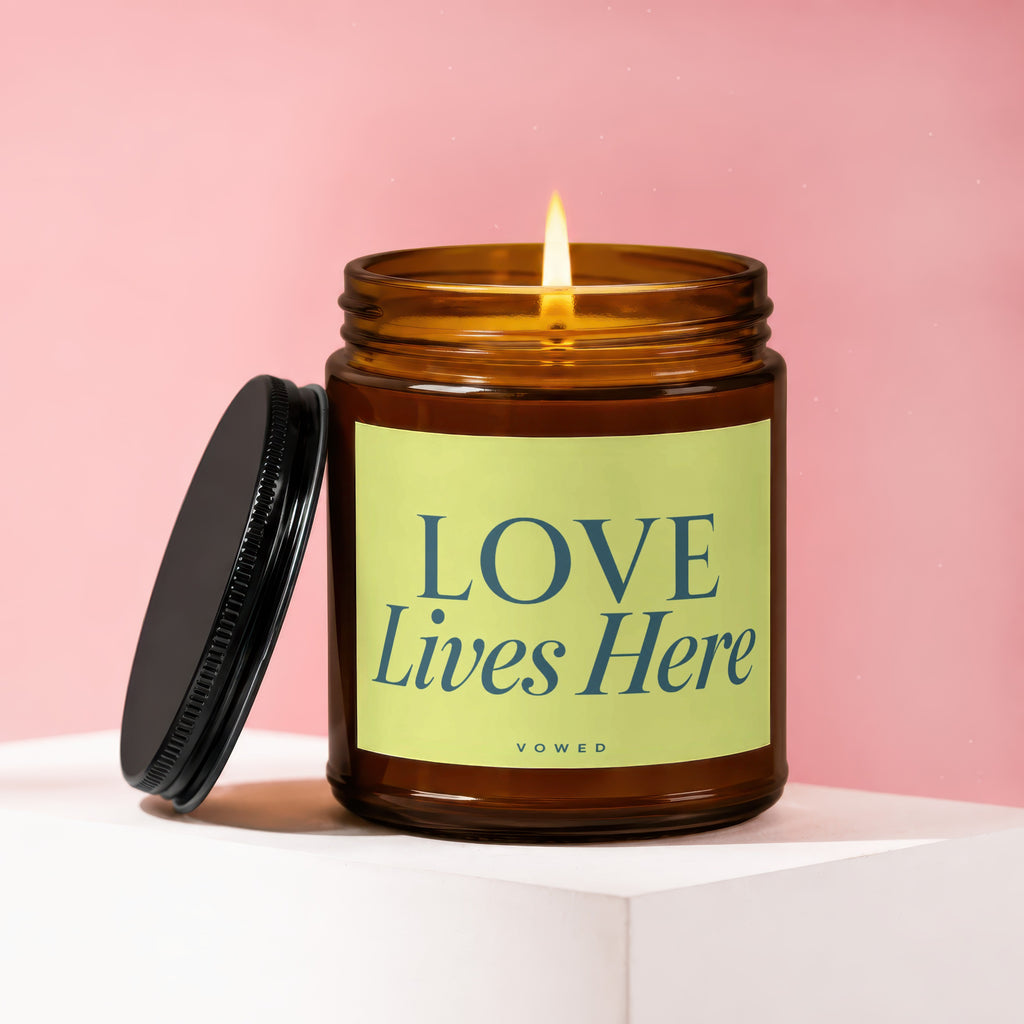 Lit amber glass candle with a pale green label reading "LOVE Lives Here" and "vowed".