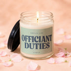 Glass jar candle with black lid, lit flame, and label reading "Smells Like Officiant Duties Vowed."