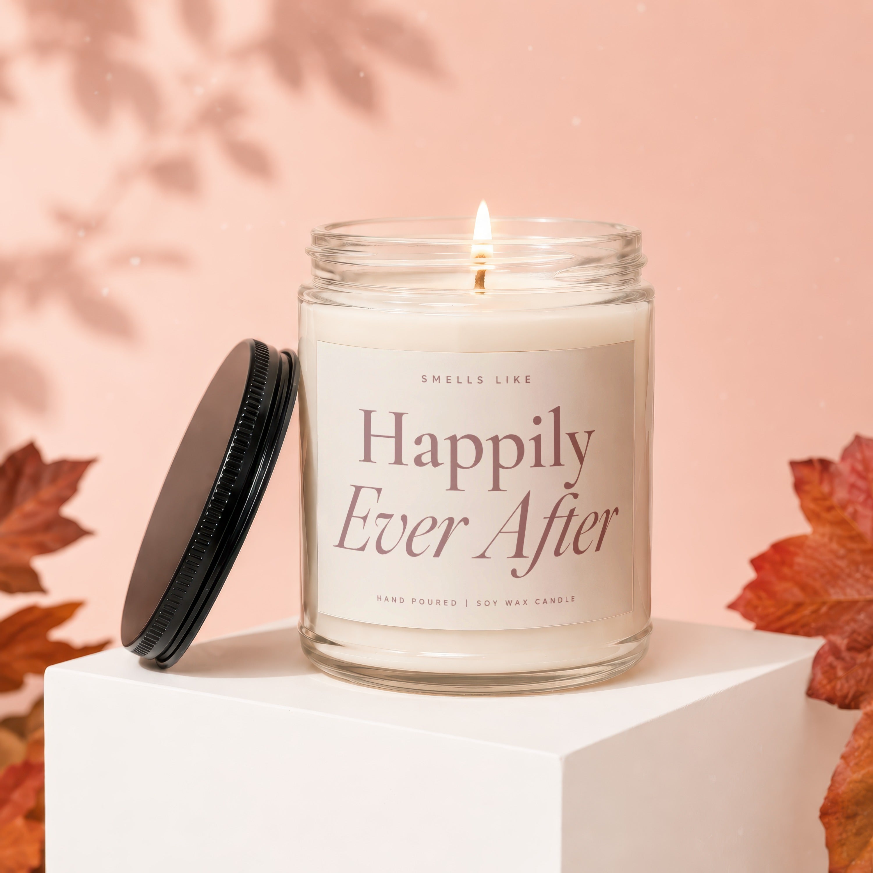 Glass jar candle labeled "Happily Ever After", lit with a dark lid beside it.