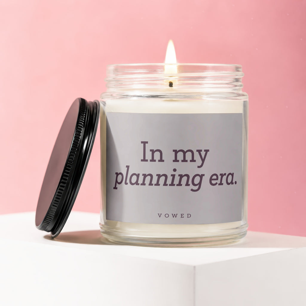 Candle with lit wick and black lid reads "In my planning era. vowed"