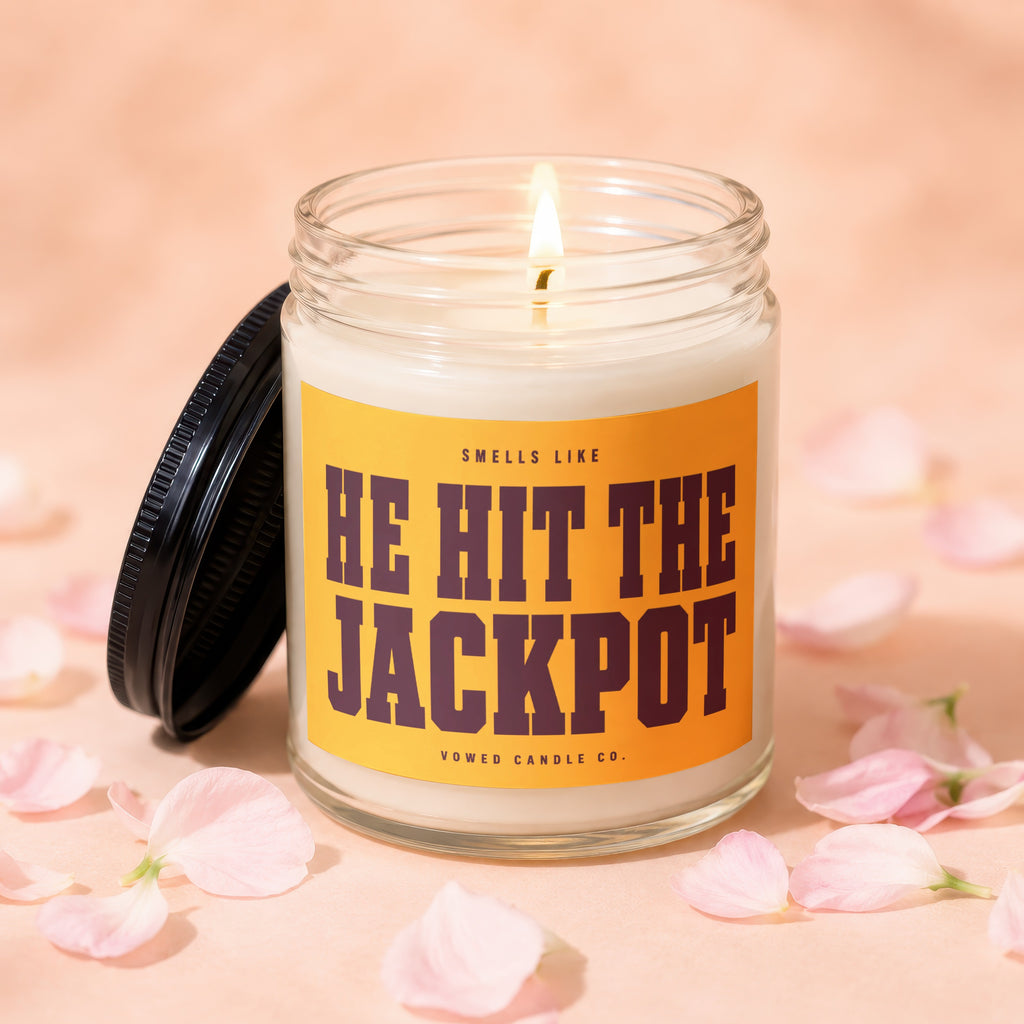 Candle with "HE HIT THE JACKPOT" label and lit wick, surrounded by pink petals.